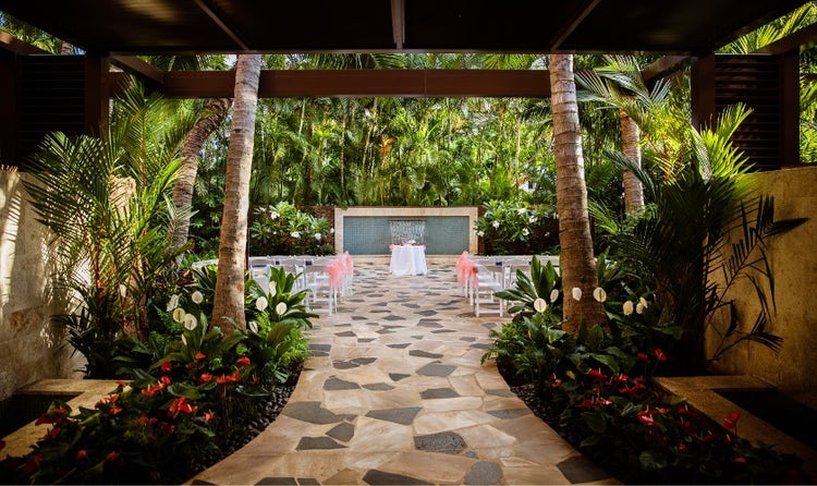 A covered, plant lined walkway leads to rows of chairs and a table in front of a fountain