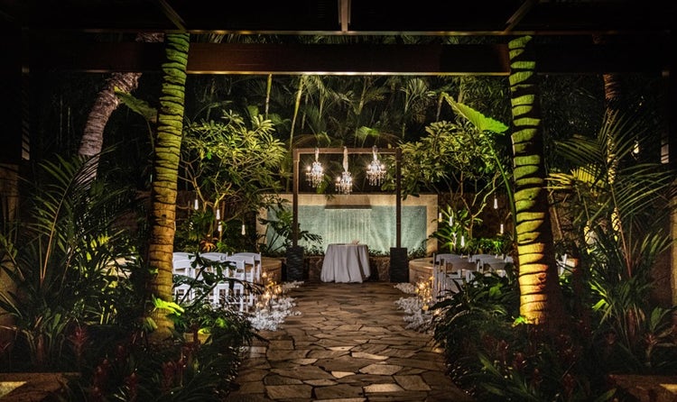 Rows of chairs and a table set up beneath a tree covered courtyard featuring chandeliers