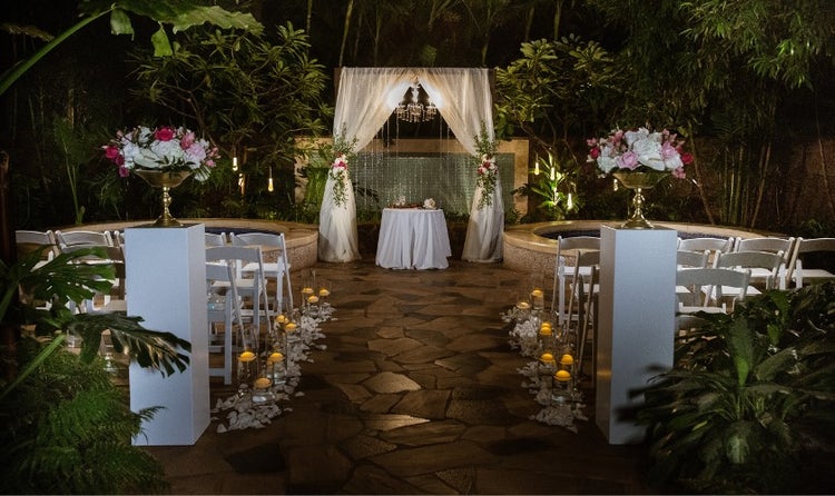 Chairs and a table under a canopy set up in front of a fountain surrounded by candles and flowers