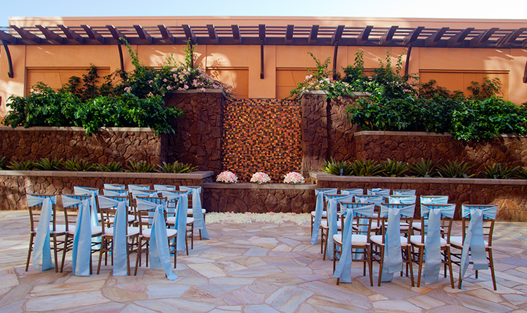 Two groups of wedding seating, each chair decorated with sashes, against a rustic, stone-wall backdrop
