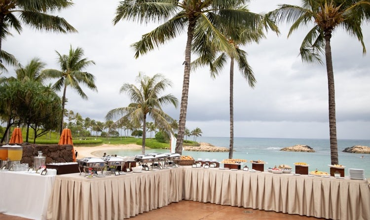 A buffet table set up filled with plates and trays in front of a palm tree lined beach