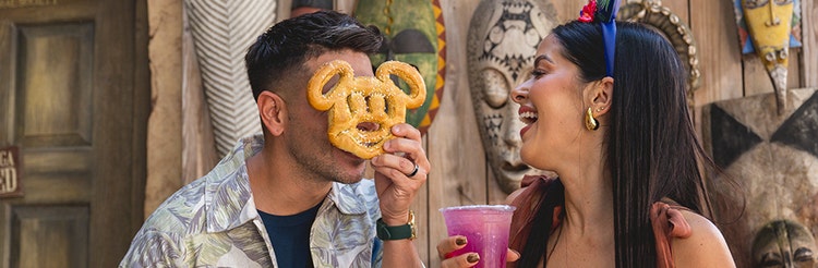 Smiling newlyweds enjoying beverages while he holds a Mickey Mouse shaped pretzel in front of his face at Disney's Animal Kingdom theme park