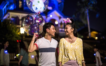 Two newlyweds smiling and holding Mickey balloons in front of Mystic Manor at Hong Kong Disneyland Resort on their honeymoon
