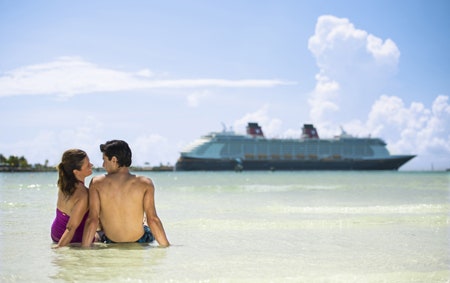 A couple sitting together in the shallow waters of a Bahamian beach with a Disney Cruise Line ship in the distance