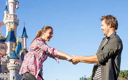 Two newlyweds laughing and holding each other's outstretched arms in front of Sleeping Beauty Castle at Disneyland Paris enjoying their honeymoon