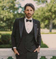 A man in a black tuxedo and bow tie smiling while sitting near floral arrangements