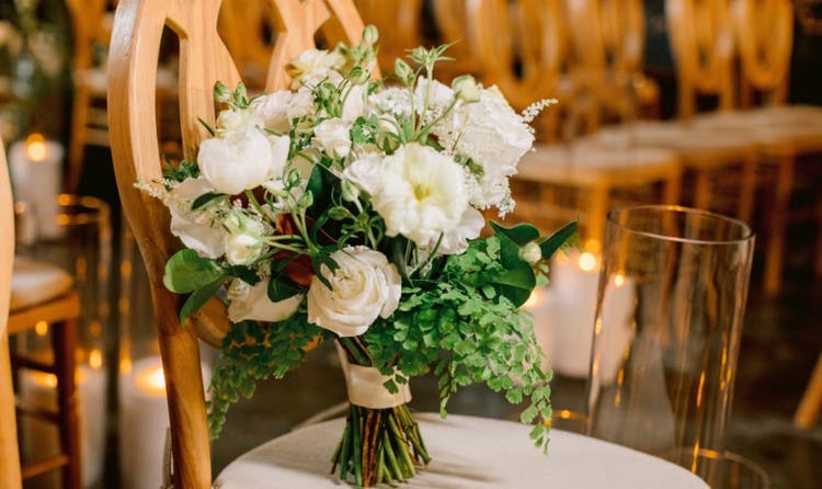 A bouquet of roses, peonies and ferns tied with ribbon, resting on a chair in a candlelit ceremony aisle