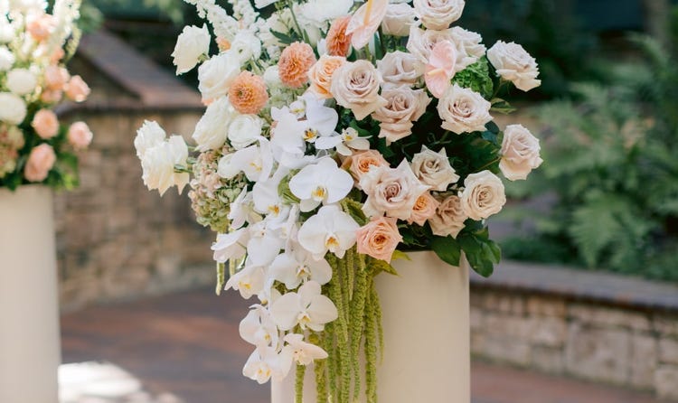 A tall flower pot overflowing with roses, orchids and trailing amaranthus at an outdoor ceremony
