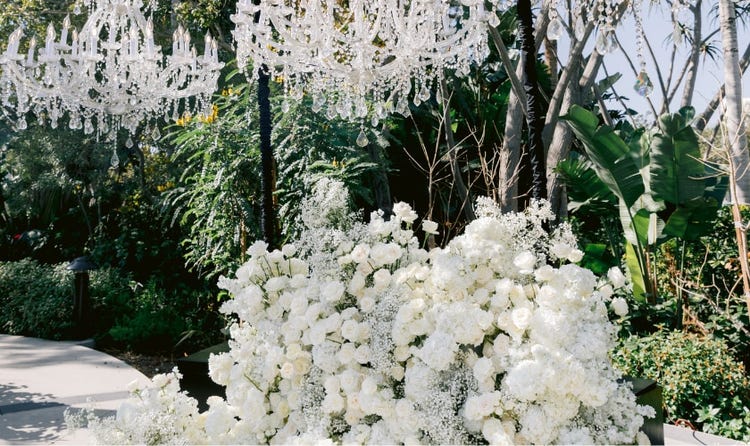 An outdoor wedding ceremony altar with oversized crystal chandeliers suspended above a wall of roses and hydrangeas