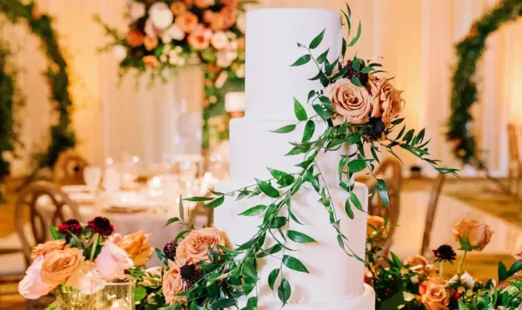 A 4 tier wedding cake draped in trailing leaves and roses, surrounded by candles and flowers in an elegant ballroom