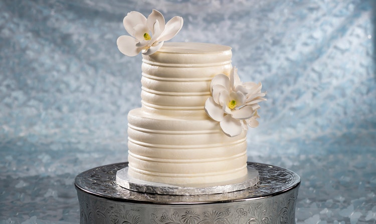A 2 tier wedding cake adorned with horizontal lines of frosting and 2 gardenia flowers