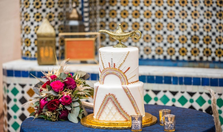 A 2 tiered cake with a sunburst design and topped with a genie’s lamp next to flowers and tea light candles