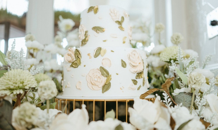 A 2-tier wedding cake with flower and leaf decor on a stand surrounded by foliage