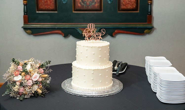 Wedding cake with a dotted bead pattern and an 'Adventure Awaits' topper, displayed beside a floral bouquet and a set of plates
