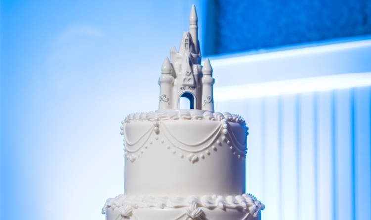 A 3 tier cake with a castle topper and intricate piped frosting next to bouquets of flowers
