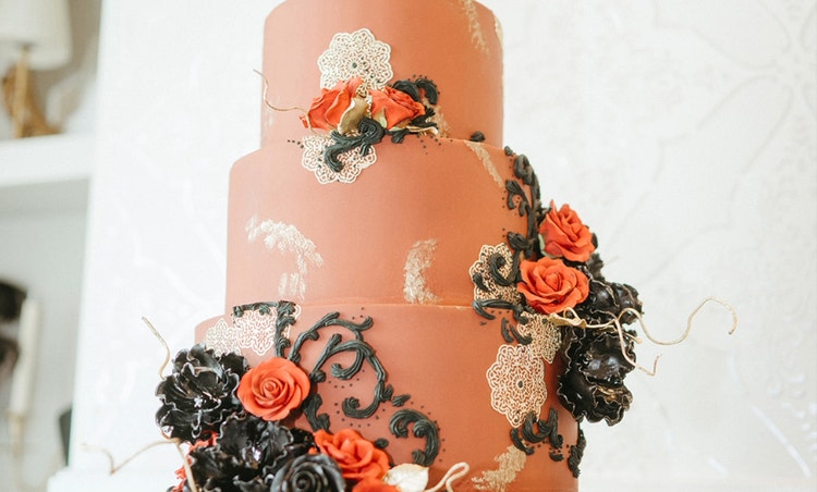 A tall multi tiered cake decorated with frosting roses and swirling vine designs sitting on an ornate cake stand