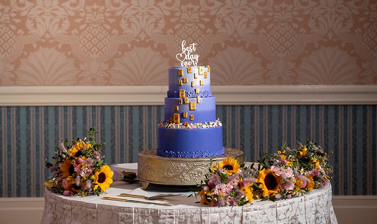 Wedding cake with a 'best day ever' topper, decorated with cascading edible tiles featuring sun designs, on a table surrounded by bouquets of flowers