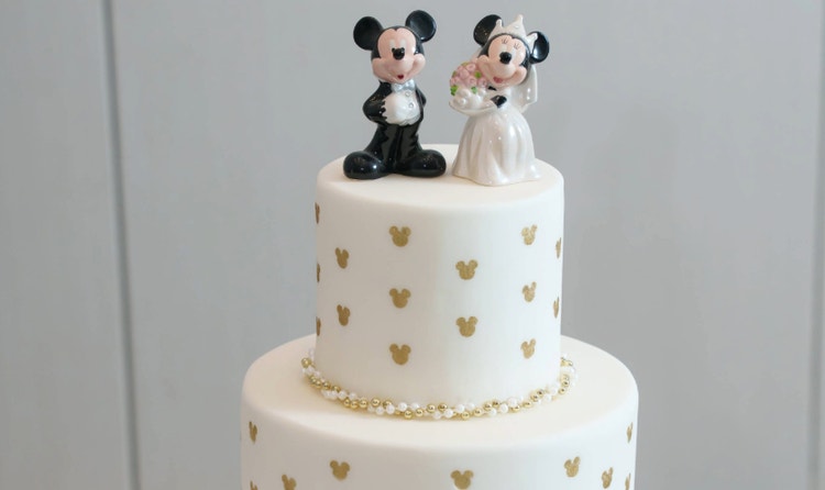 A stacked wedding cake with a Mickey and Minnie wedding topper, Micky Mouse ear patterns and beaded trim on an ornate stand