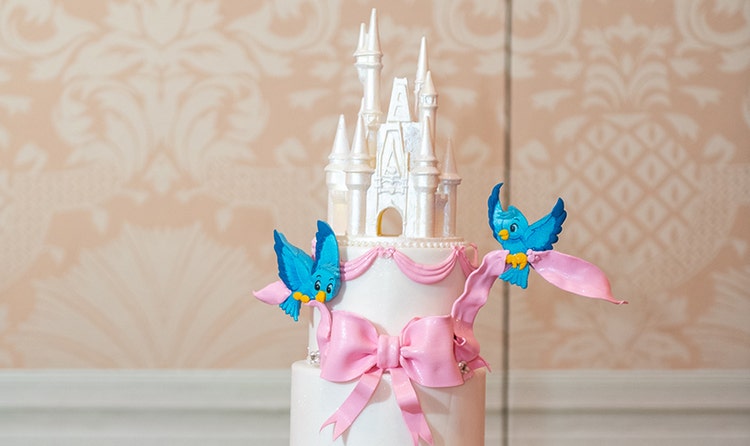 Wedding cake with Cinderella themed decorations featuring a Cinderella Castle topper, bluebirds with ribbons, a carriage and more