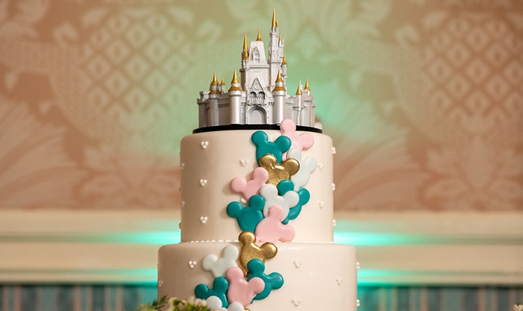 Wedding cake with a Cinderella Castle topper, cascading Mickey shaped accents and a beaded heart pattern, on a table with flower bouquets
