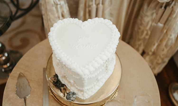 A heart shaped cake with ruffled frosting and ‘Just Married’ written on top, and a knife, server, and dog and cat figurines placed nearby